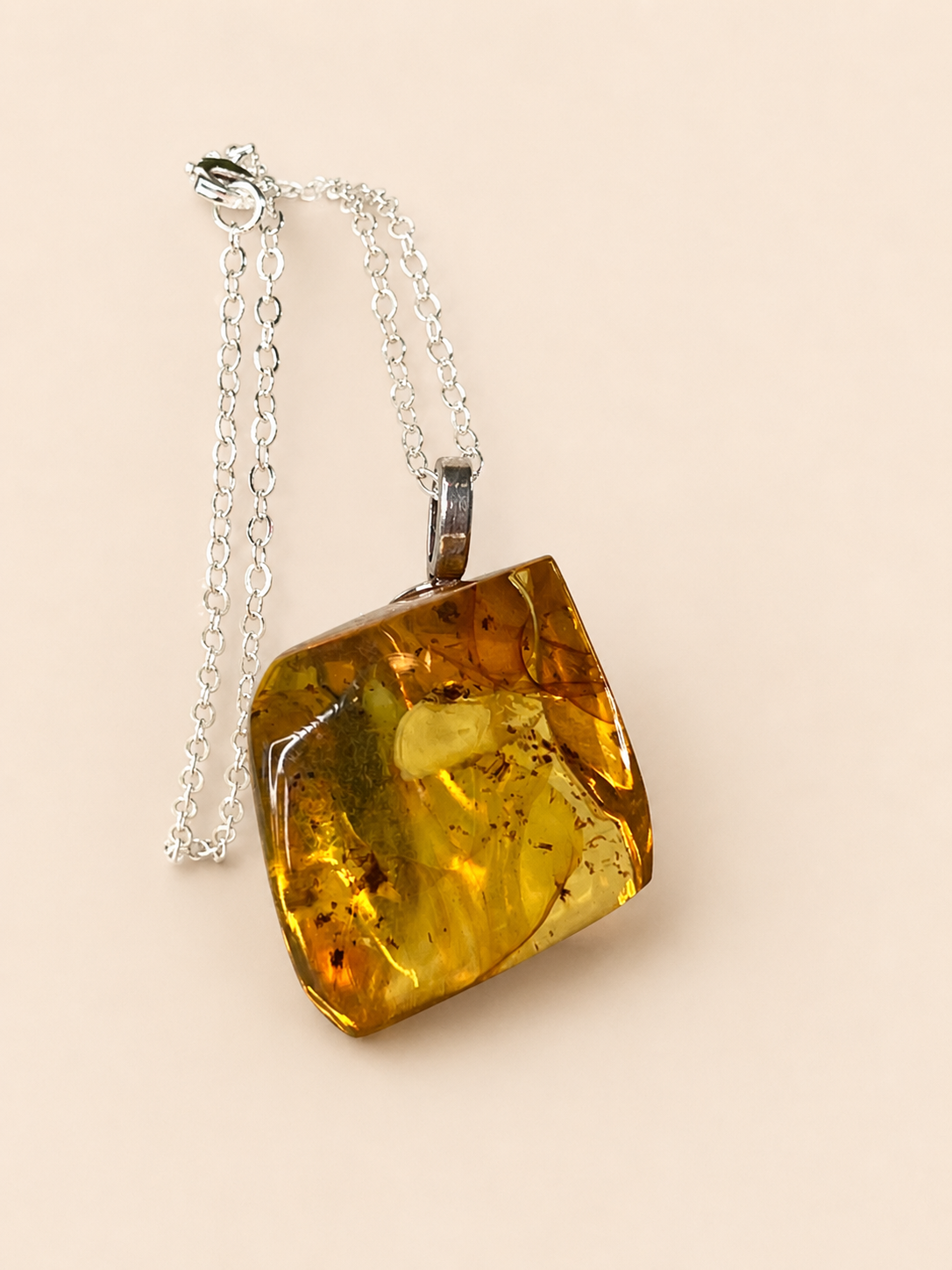 A chunky, irregular-shaped amber pendant necklace with a silver chain, displayed on a solid light beige background.