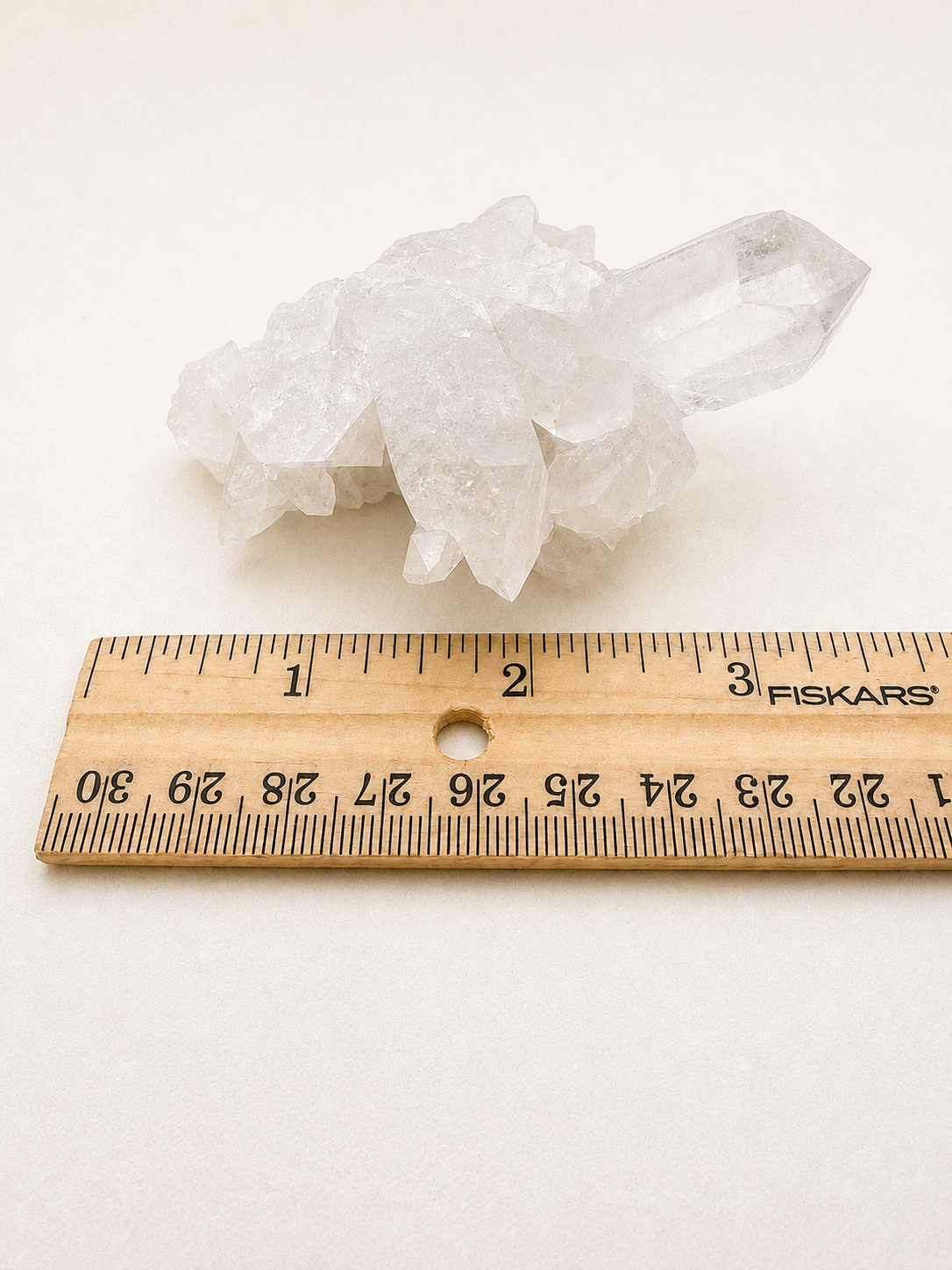 A cluster of clear quartz crystals sits on a plain white surface above a wooden Fiskars ruler, showing their size comparison.