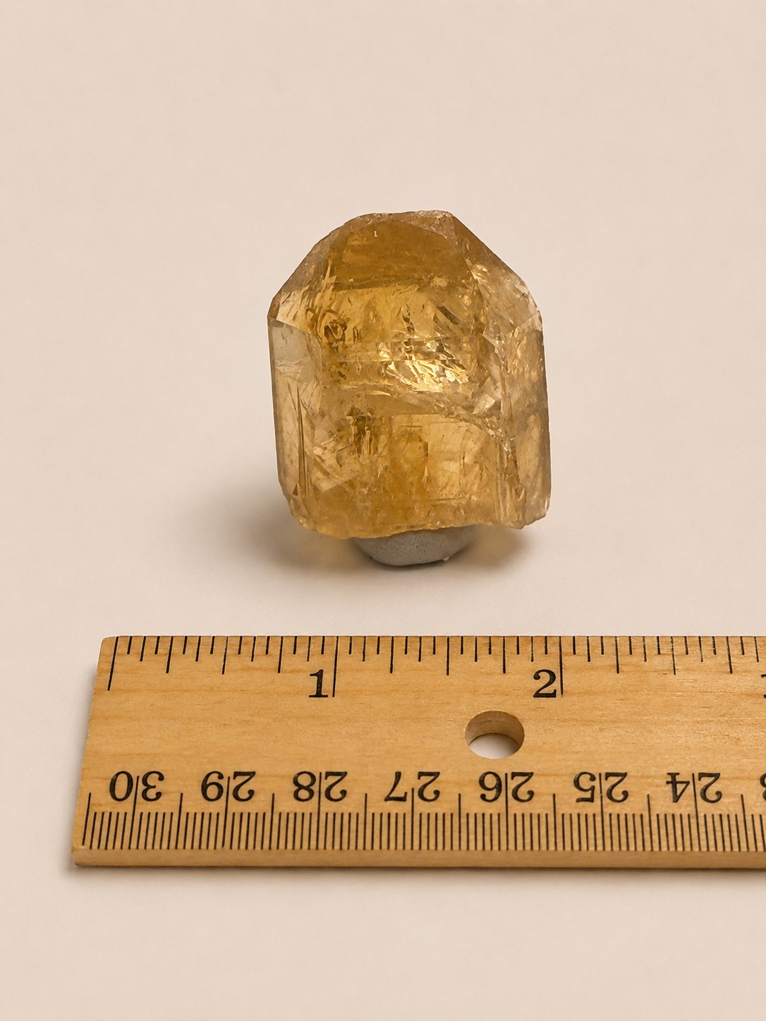 A small translucent yellow crystal or gemstone with natural faceted edges, photographed on a plain light beige background above a wooden ruler for scale.