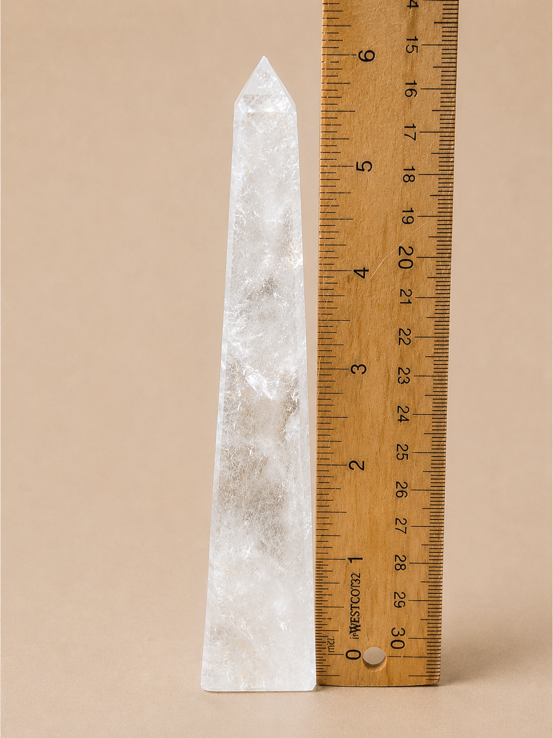 A tall, pointed clear quartz crystal obelisk shown next to a wooden ruler with inch markings on a solid beige background.