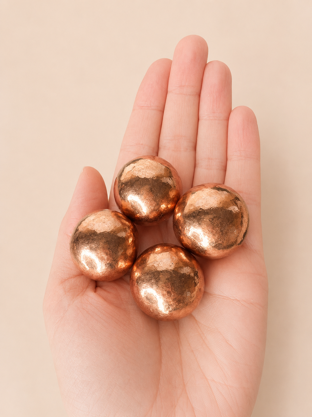 Four metallic copper spheres resting in the palm of a hand against a solid beige background.