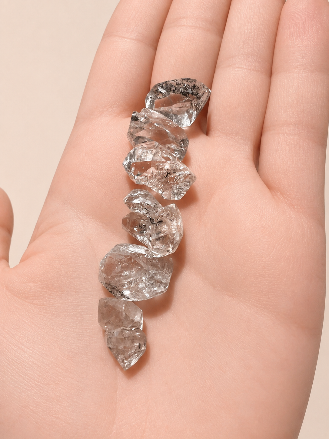 Five rough, uncut clear gemstones held on the back of an open hand against a soft beige background.