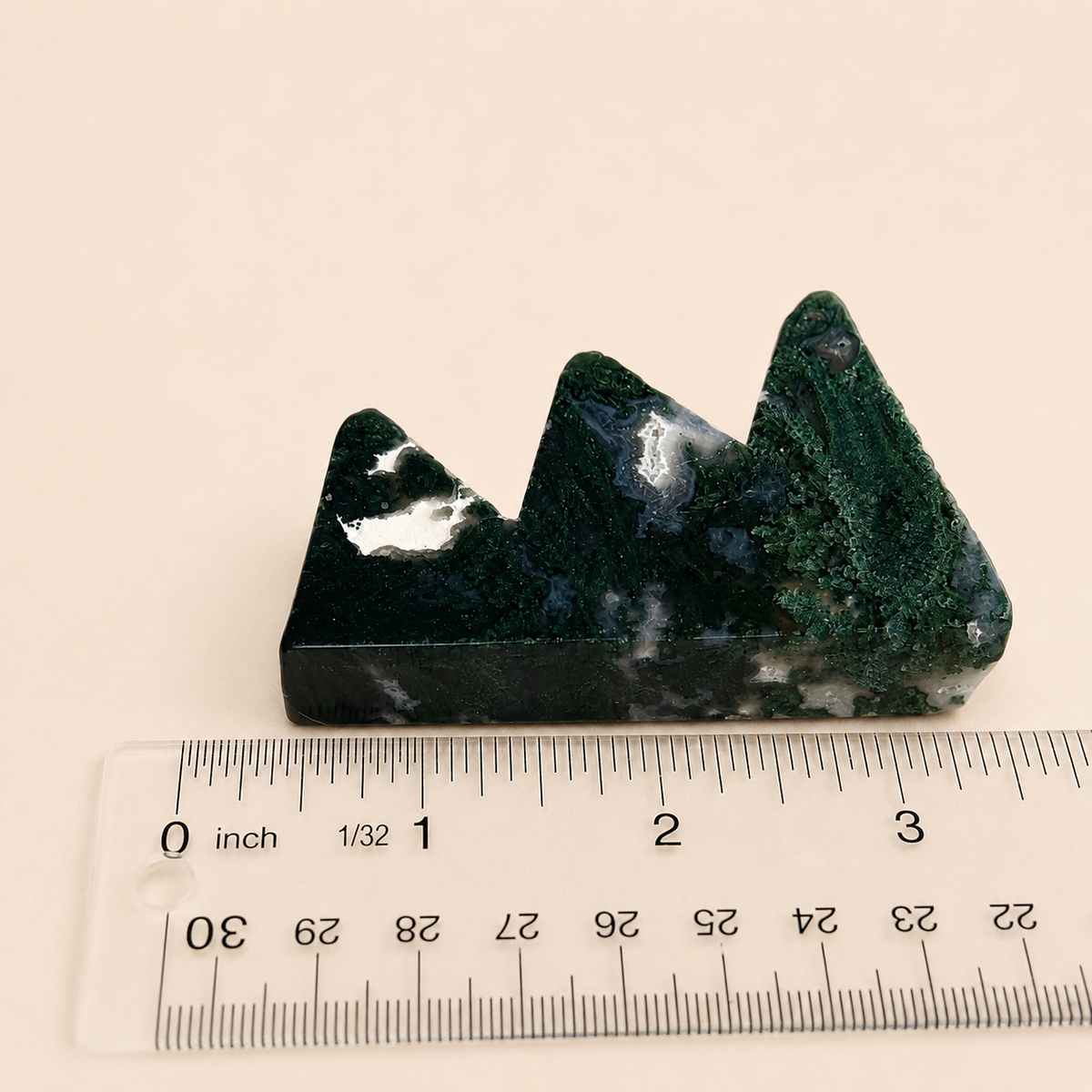 A small mountain-shaped green stone or crystal cluster placed on a clear ruler against a plain light beige background.
