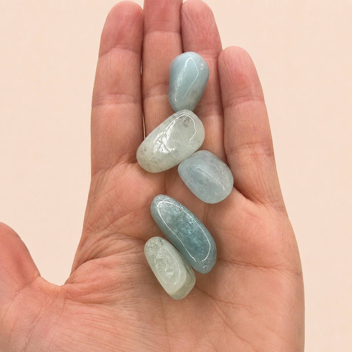Five polished light blue tumbled stones resting on an open human hand against a soft beige background.