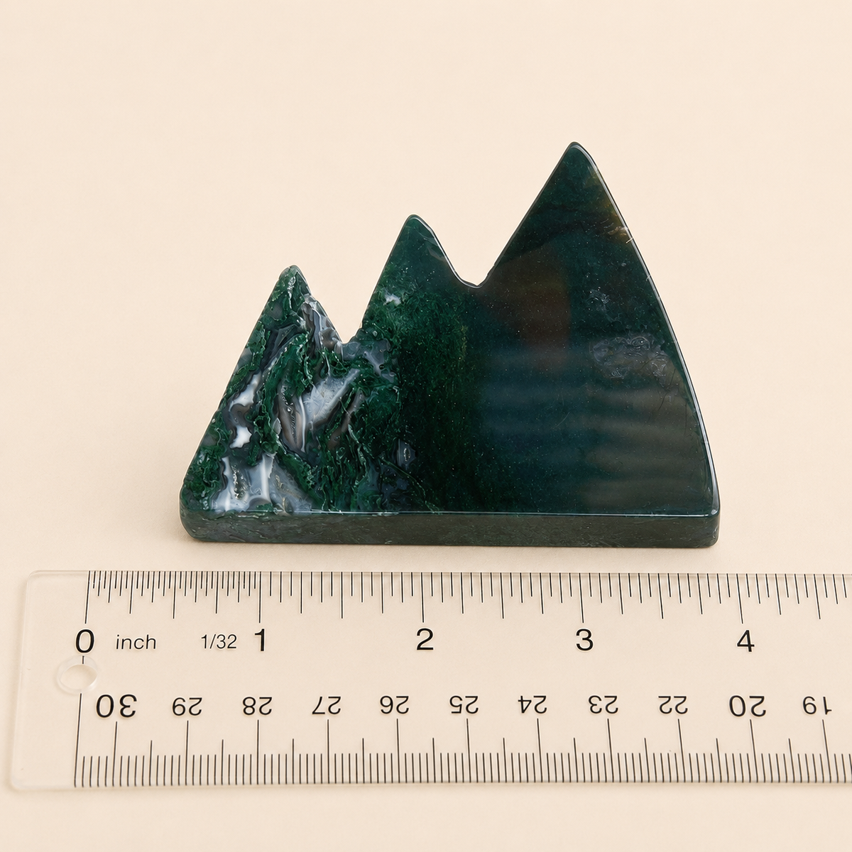 A small mountain-shaped dark green polished stone sculpture shown above a clear ruler on a light beige background.