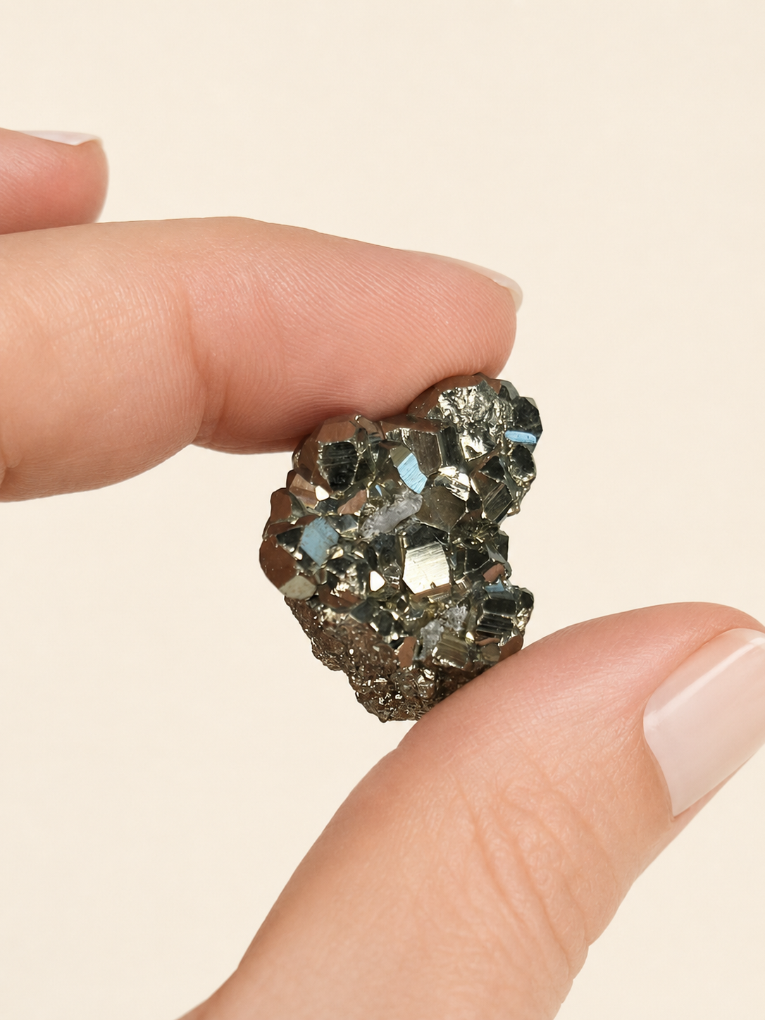 A close-up of a hand holding a small, rough, iridescent crystal cluster or gemstone against a plain light beige background.