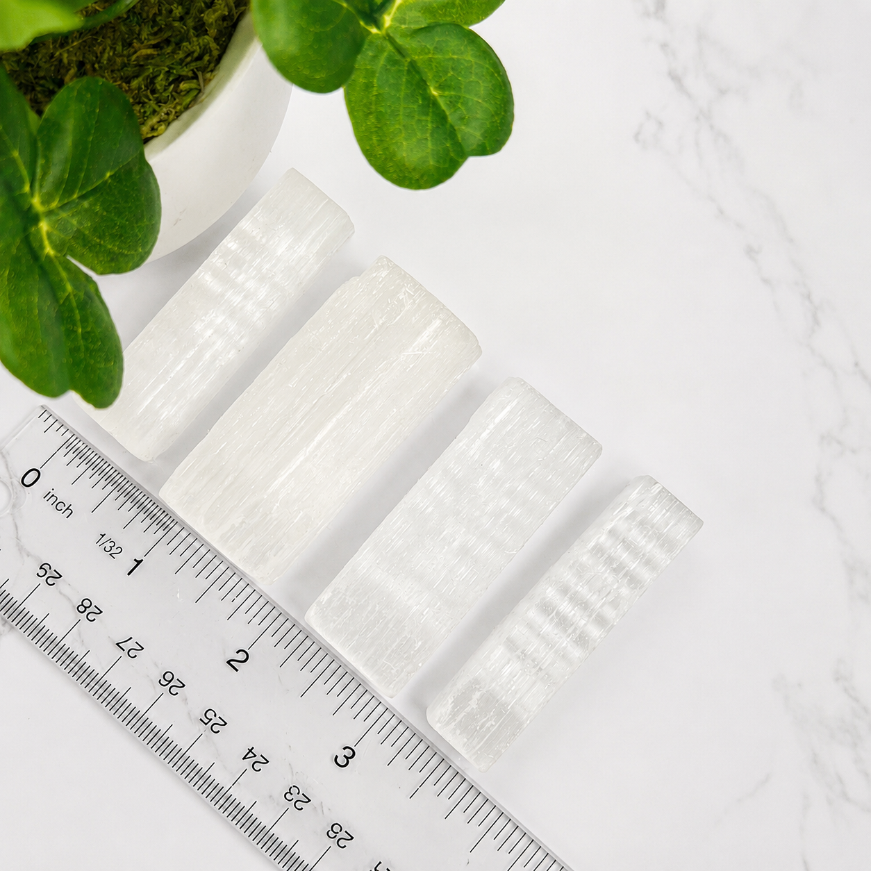 Four translucent white rectangular crystal selenite or stone pieces arranged diagonally on a white marble surface, with a ruler and green plant leaves visible for scale and styling.