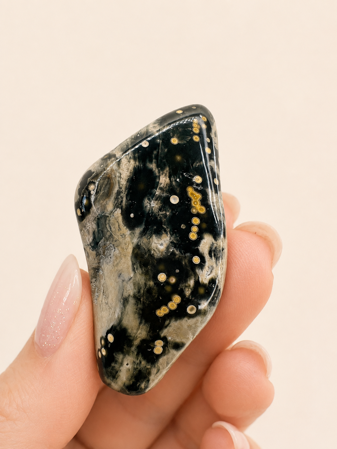 A polished black tumbled gemstone with golden and white star-like speckles, held in a person's hand against a soft beige background.