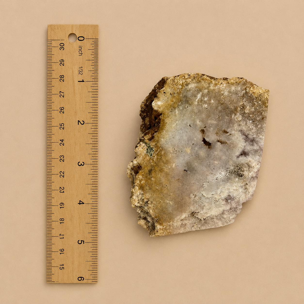 A rough mineral or geode specimen with a pale gray to off-white crystalline center and tan to brown outer matrix, shown next to a wooden ruler on a plain beige background.