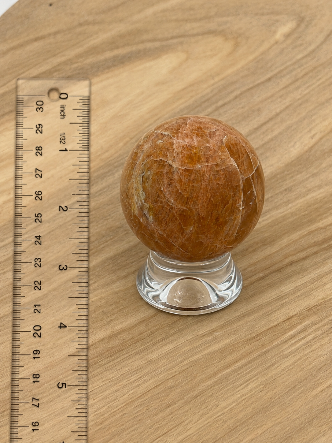 A polished orange-brown crystal sphere on a clear glass stand, shown next to a wooden ruler on a light wood grain surface.