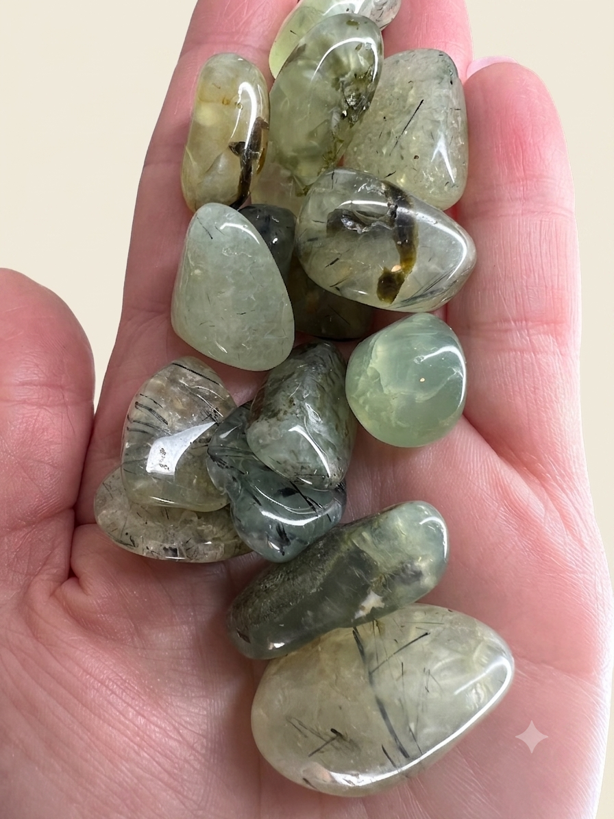 A hand holding a cluster of polished tumbled gemstones in various shades of green, gray, and beige, showing their smooth, rounded shapes and natural inclusions.