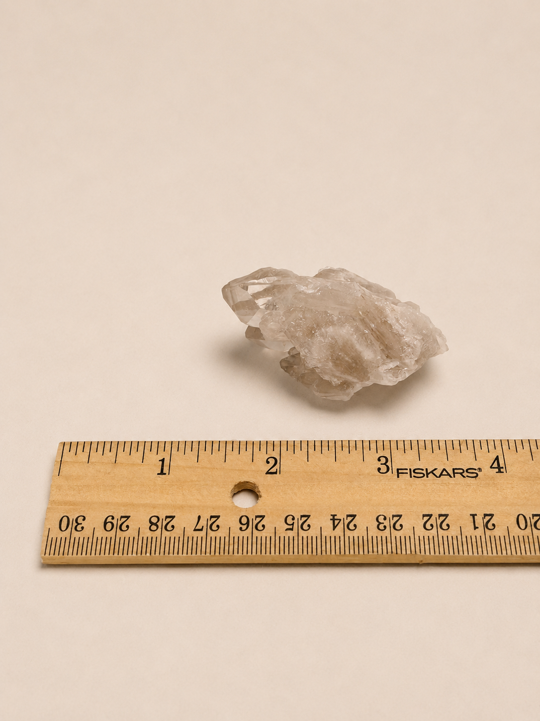 A small translucent pale crystal or raw gemstone placed above a wooden ruler on a plain warm beige background. The ruler shows centimeter markings and the brand name FISKARS.