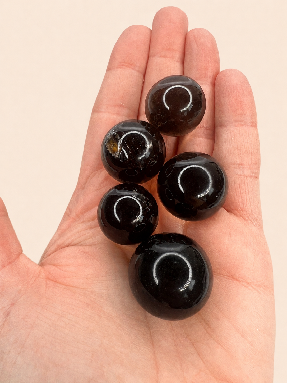 Five polished black smoky quartz spehres resting on an open hand, photographed against a plain light beige background.