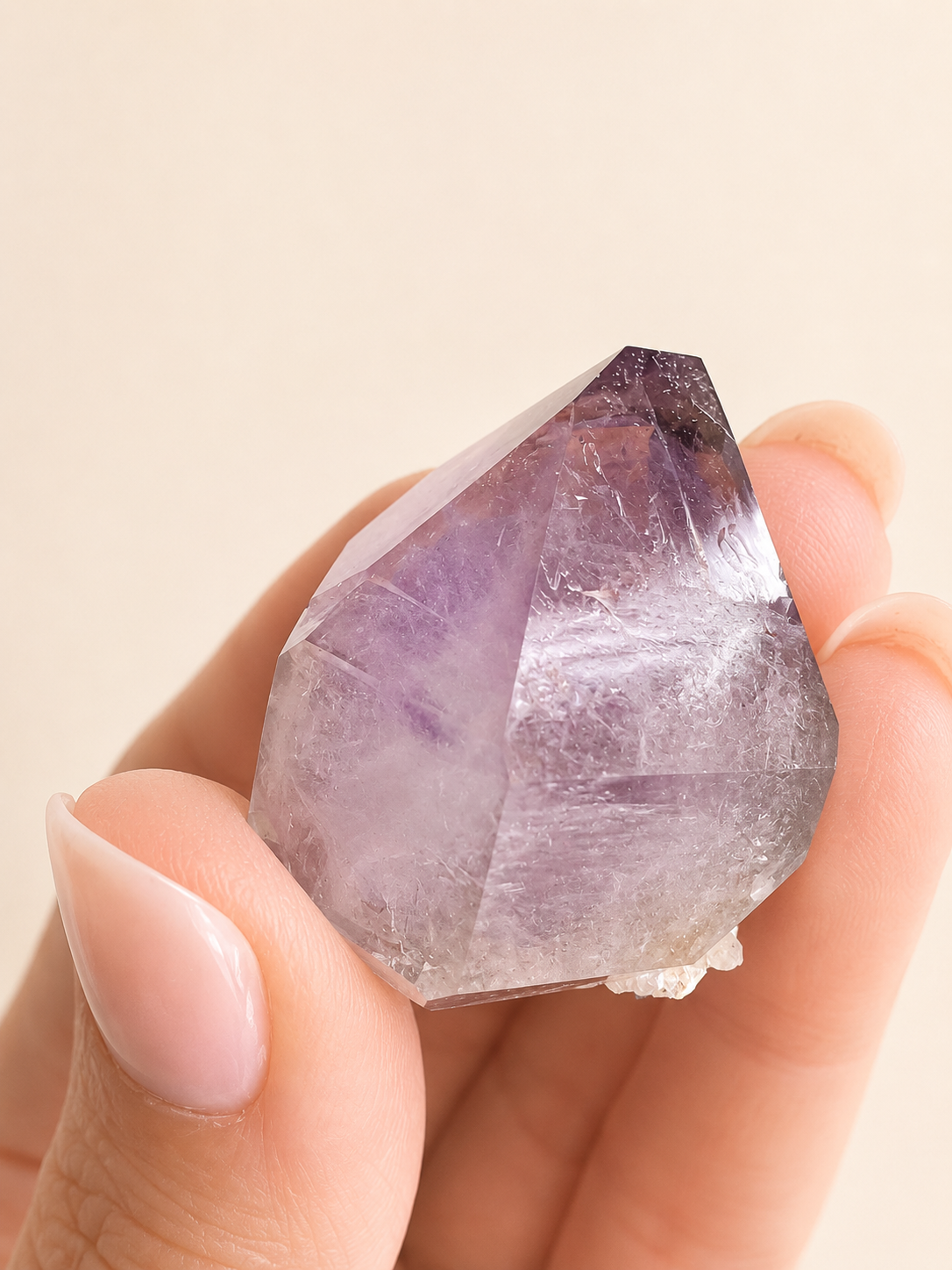 A hand with pale pink manicured nails holds a faceted purple crystal point against a soft beige background.