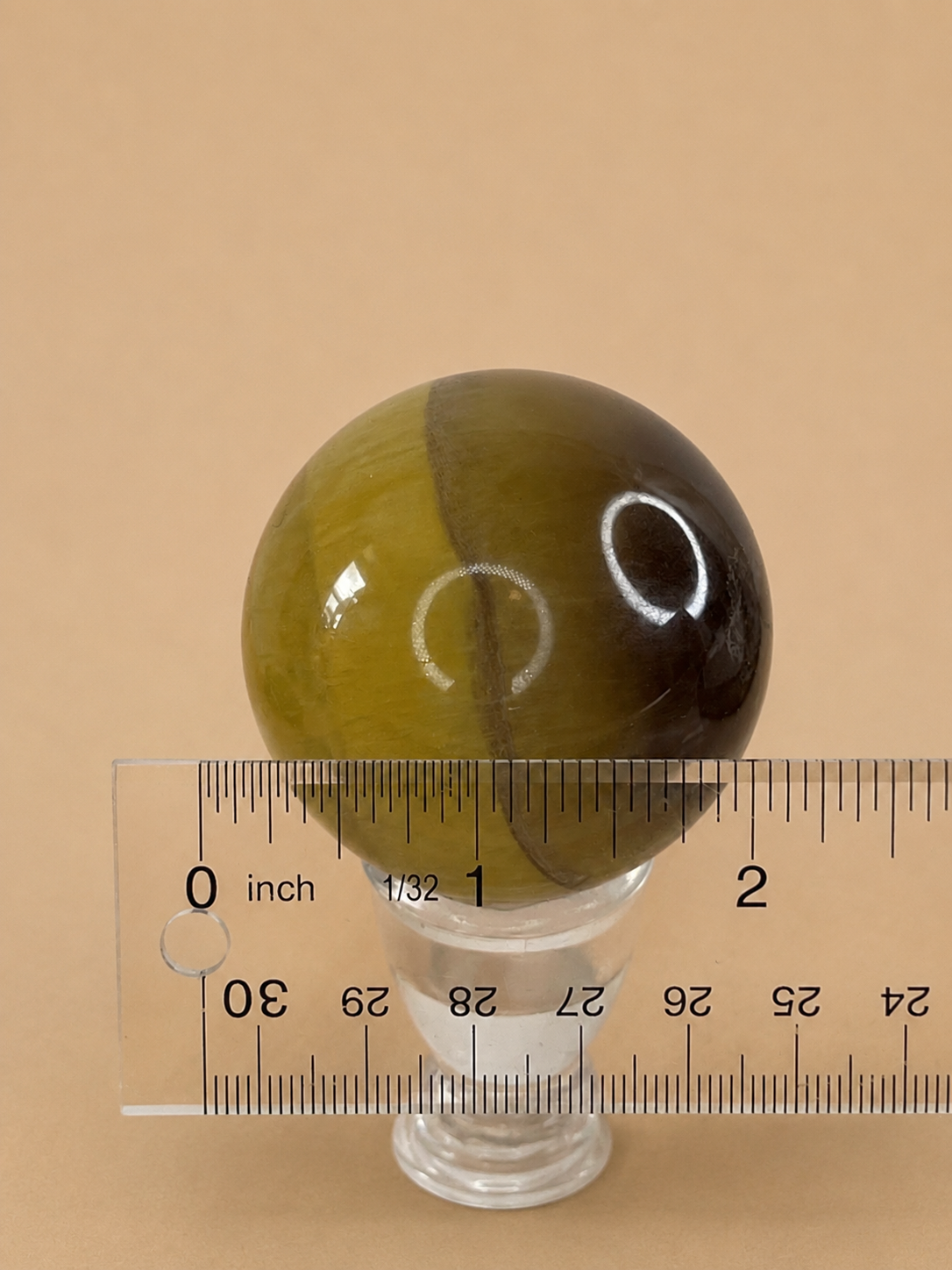 A polished oval stone ornament in olive green and dark brown, displayed on a clear glass stand with a clear plastic ruler below for scale, set against a solid beige background.