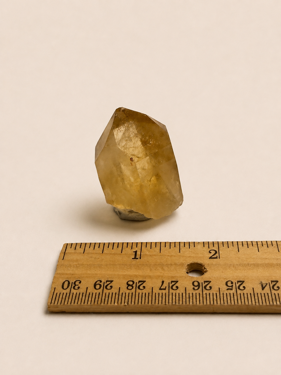 A small rough translucent yellow crystal or stone standing upright on a white background, with a wooden ruler below it for scale.