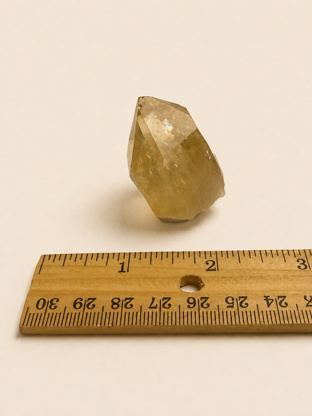 A small faceted yellow crystal or stone placed above a wooden ruler on a plain light beige background.