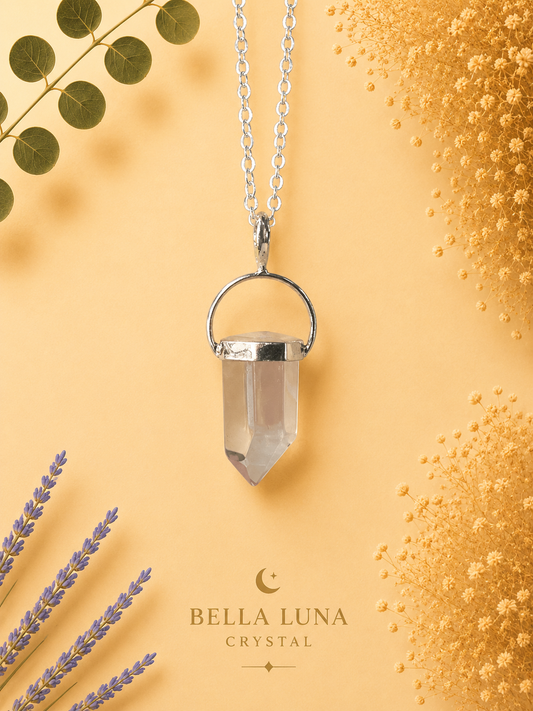 A silver-toned pendant necklace featuring a clear crystal point hanging from a delicate chain, styled on a warm peach background with dried lavender, eucalyptus leaves, and textured floral elements. The brand name 'BELLA LUNA CRYSTAL' is printed at the bottom.