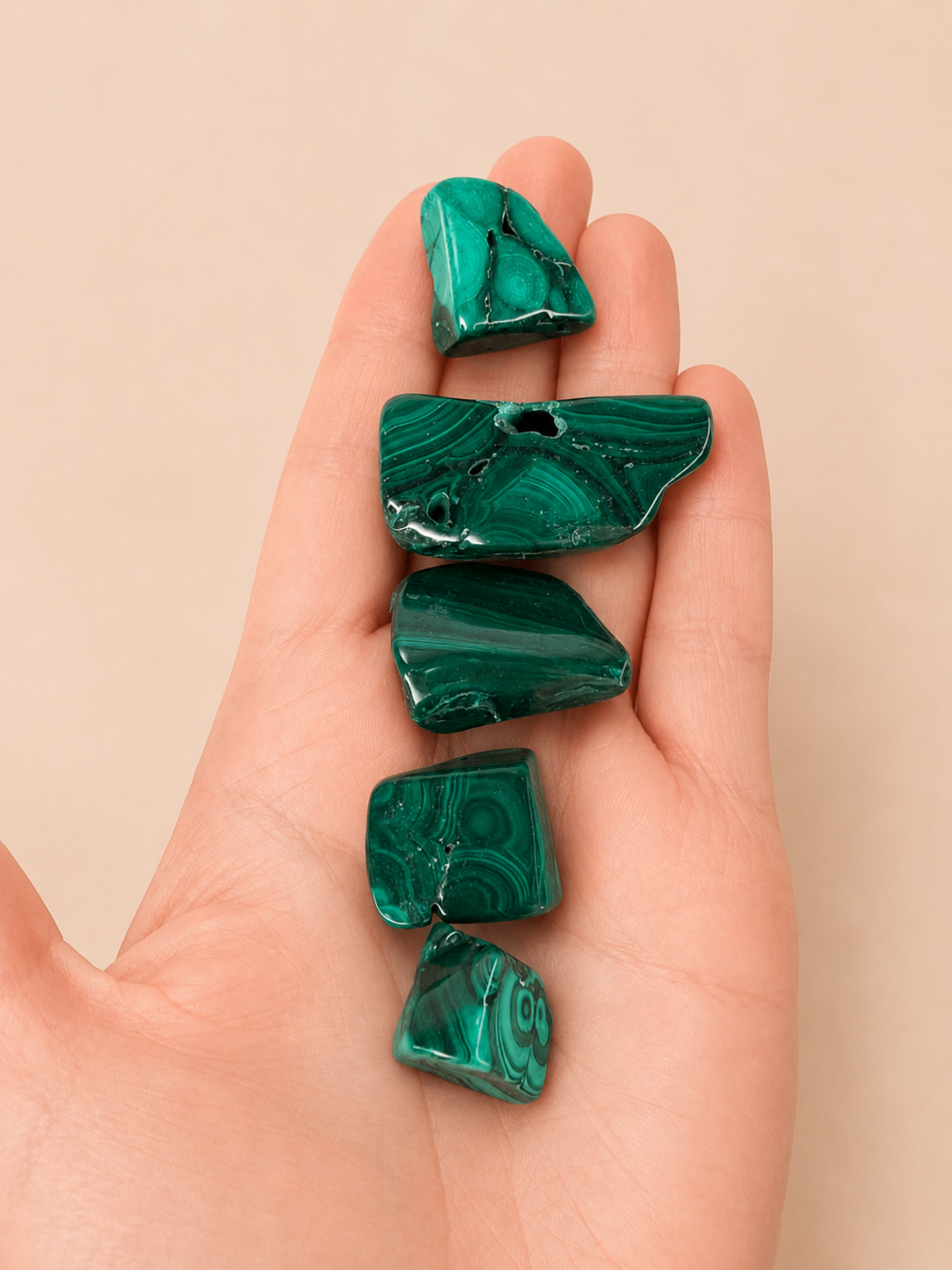 Five polished green malachite tumbled stone beads with natural banding, held in the palm of a hand against a solid beige background.