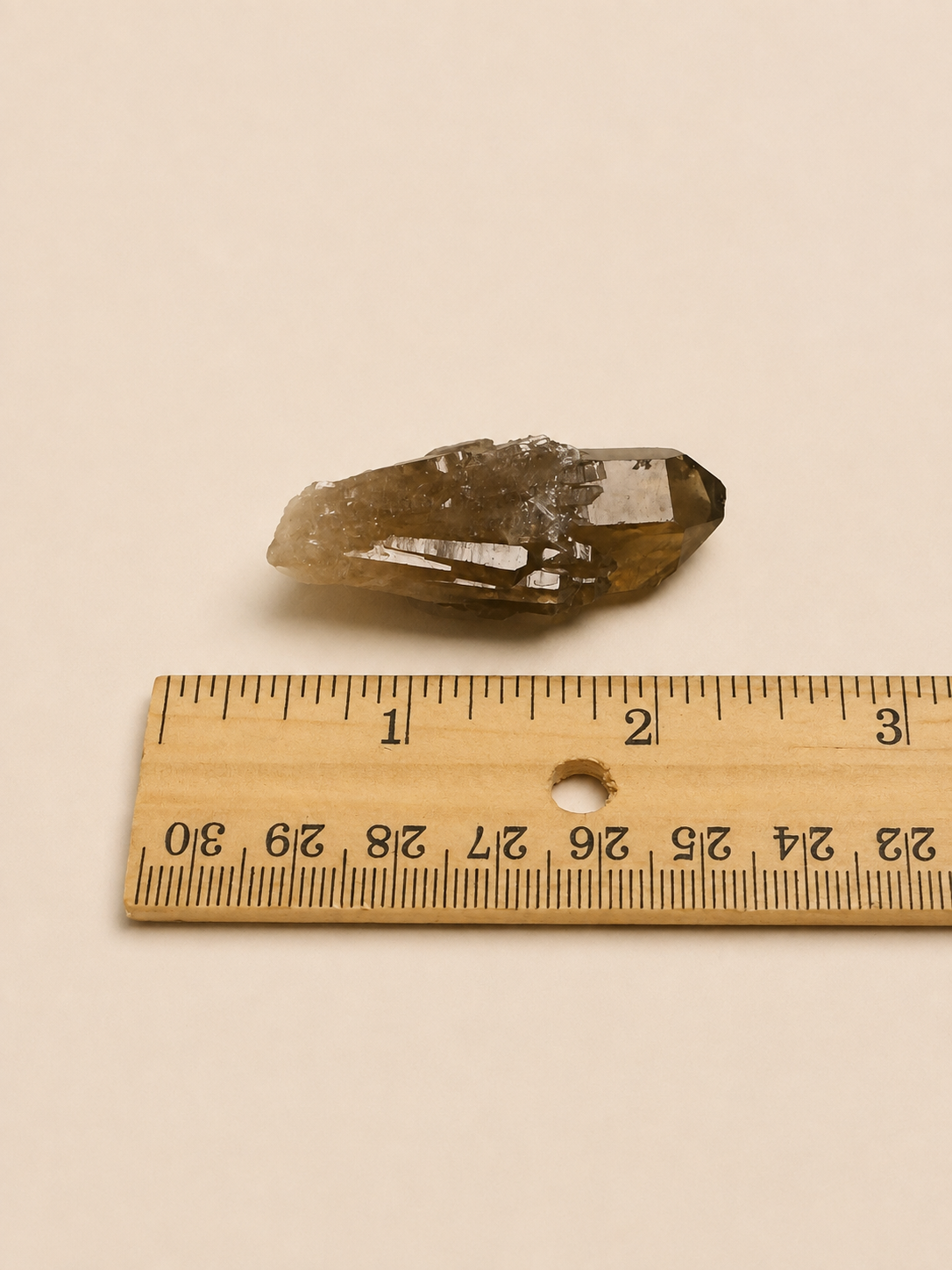 A small natural smoky quartz crystal point shown next to a wooden ruler on a plain beige background.