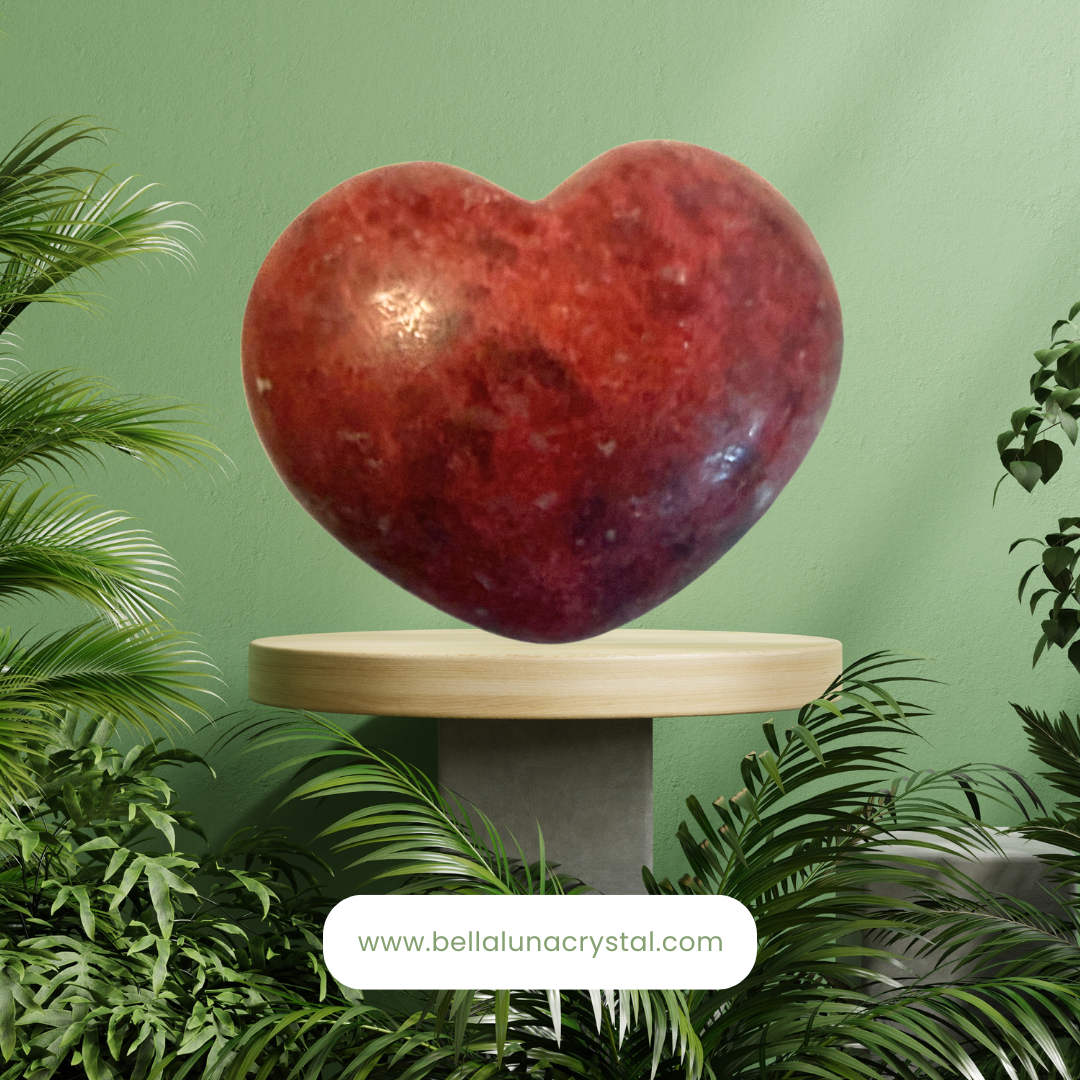 Heart-shaped crystal on a pedestal with green plants around, brand 'Bella Luna Crystal' visible.