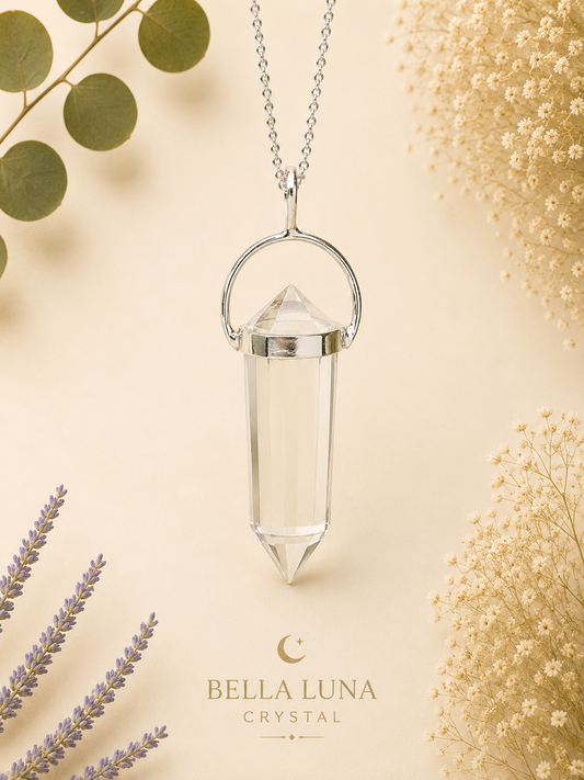 A silver necklace with a clear crystal point pendant, displayed on a warm beige background with decorative dried flowers and greenery. The brand name 'BELLA LUNA CRYSTAL' is printed at the bottom.