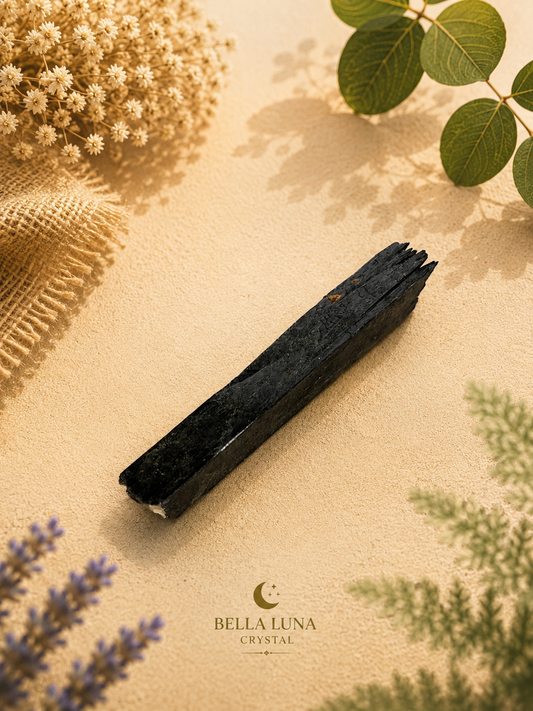 A dark polished crystal rod lies diagonally on a textured beige surface, surrounded by dried flowers, green leaves, and burlap. The Bella Luna Crystal logo is printed at the bottom center.