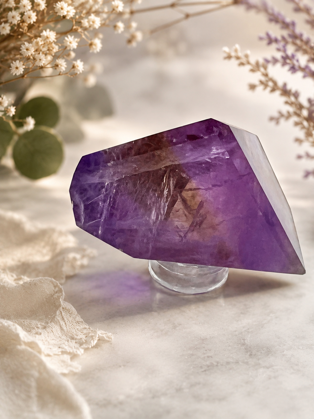 A polished purple and yellow crystal point resting on a small clear stand, photographed on a soft light surface with dried flowers and greenery around it.