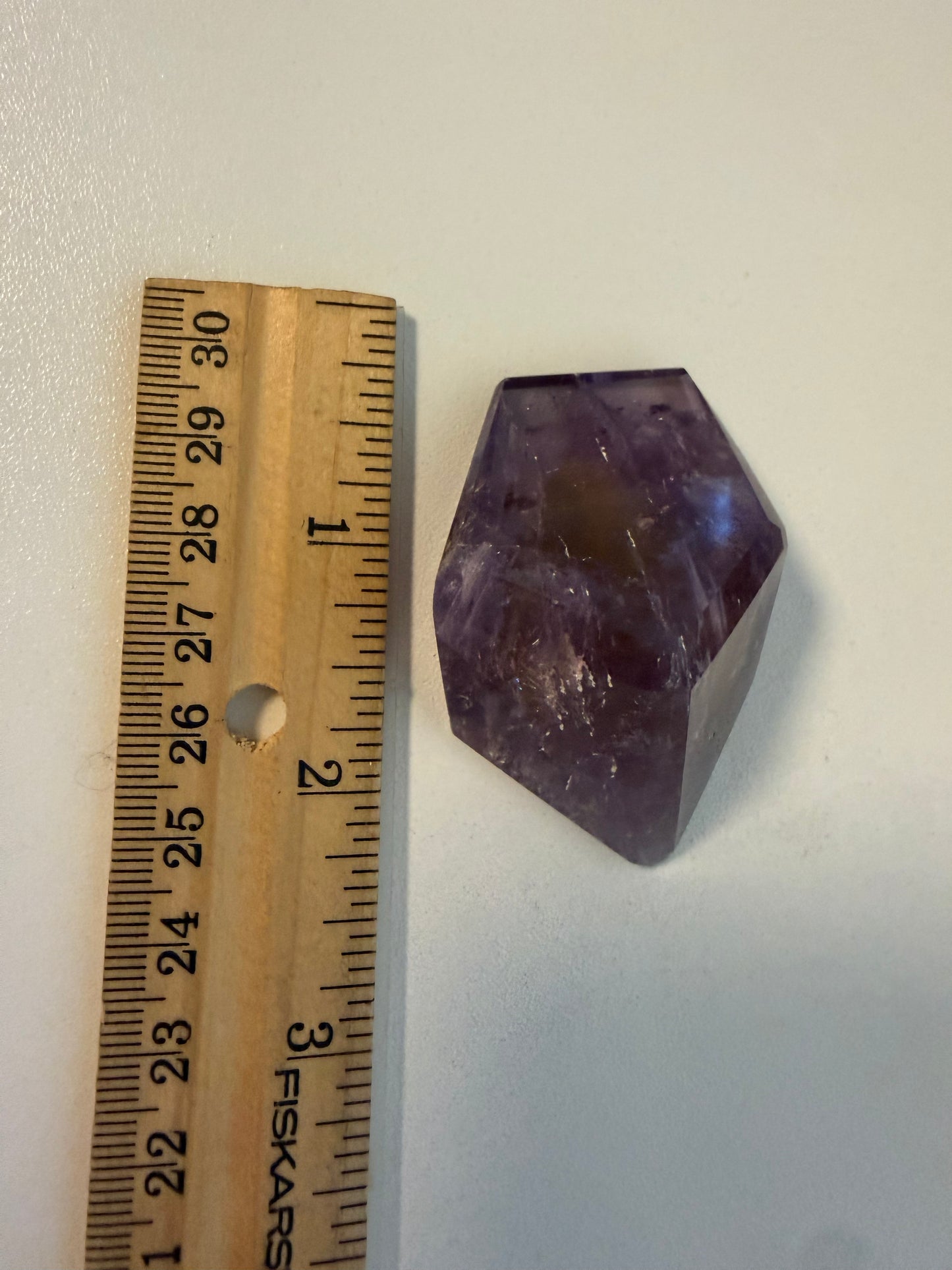 A faceted purple and yellow crystal stone placed next to a wooden ruler on a plain light background, showing its size.