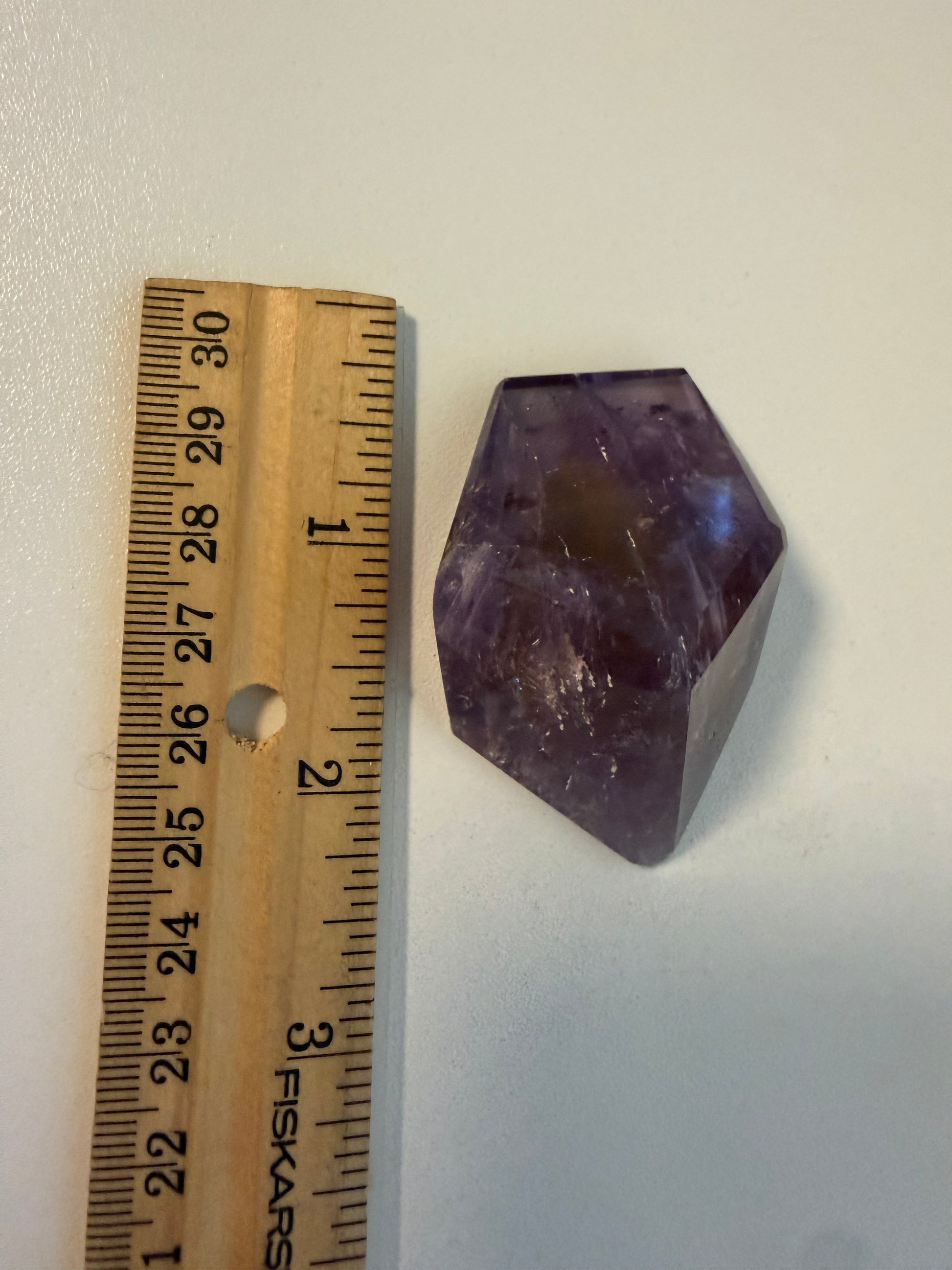 A faceted purple and yellow crystal stone placed next to a wooden ruler on a plain light background, showing its size.