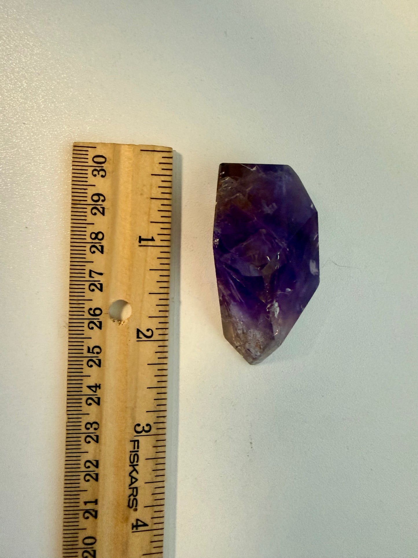 A raw purple amethyst crystal stone shown next to a wooden ruler for scale on a plain light background.
