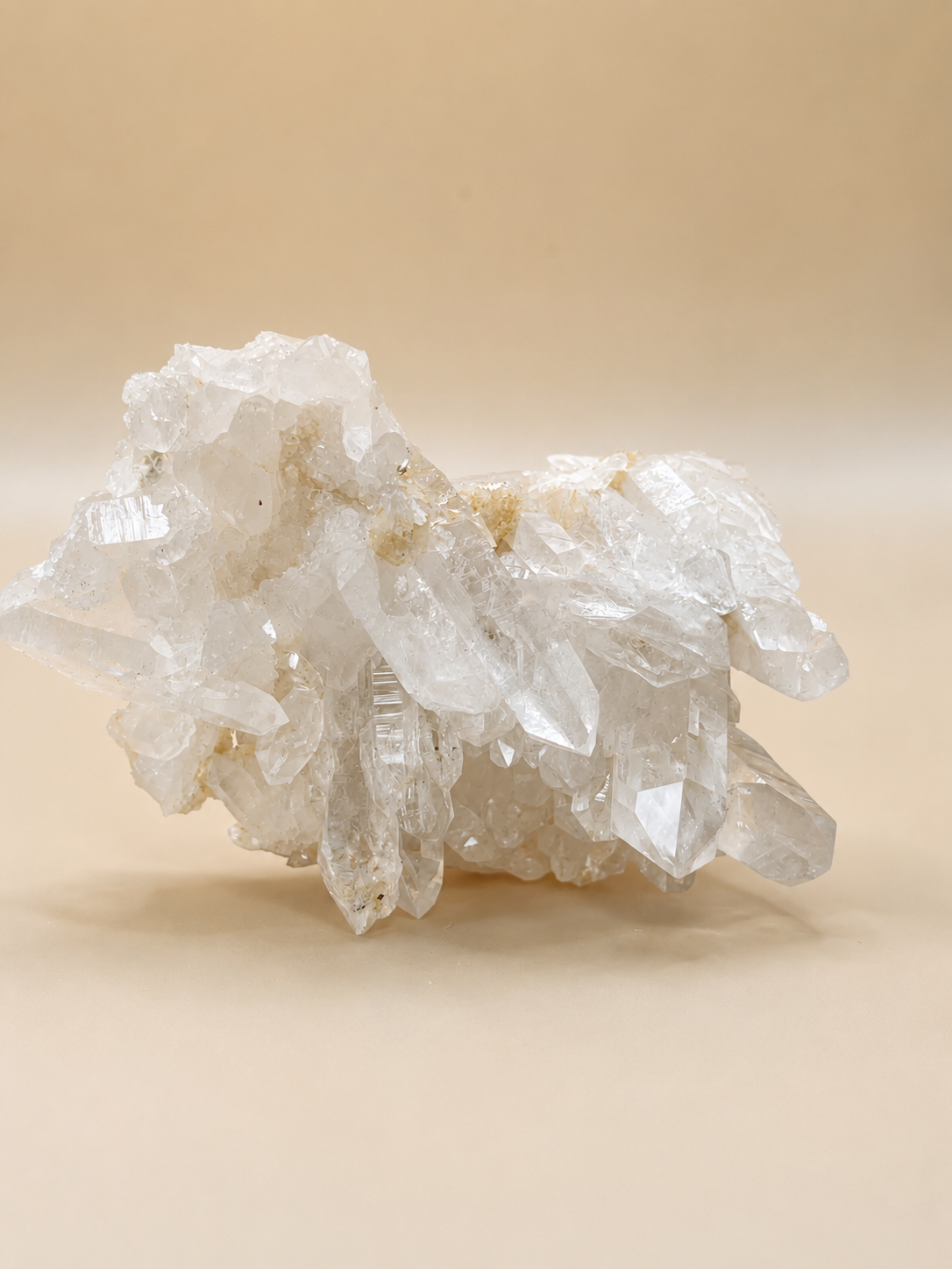 A cluster of clear quartz crystal points with natural rough edges and subtle beige mineral inclusions, displayed on a smooth beige background.