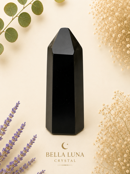 A polished black crystal point tower centered on a warm cream background, surrounded by dried flowers, green leaves, and lavender sprigs. The brand name 'BELLA LUNA CRYSTAL' appears below the crystal.