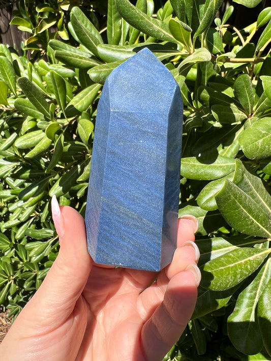 A hand holding a blue Aventurine crystal with a pyramidal shape, with greenery in the background.