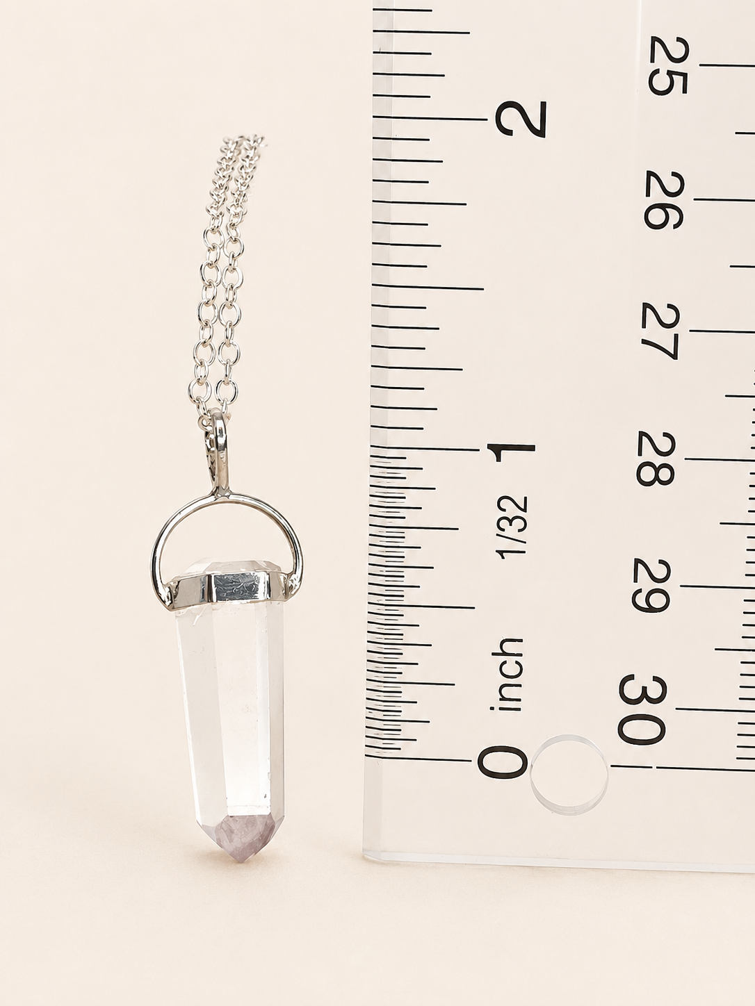A silver-toned crystal point pendant necklace on a delicate chain, shown next to a clear ruler for scale on a light beige background.