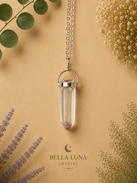 A clear crystal point pendant necklace with a silver-toned metal cap and hanger, suspended on a chain against a warm beige background with botanical props and the Bella Luna Crystal logo.