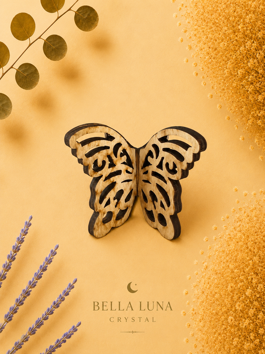 A laser-cut wooden butterfly ornament with intricate openwork patterns, displayed on a warm yellow background with decorative dried lavender, gold leaves, and textured gold elements. The brand name 'BELLA LUNA CRYSTAL' is printed at the bottom.