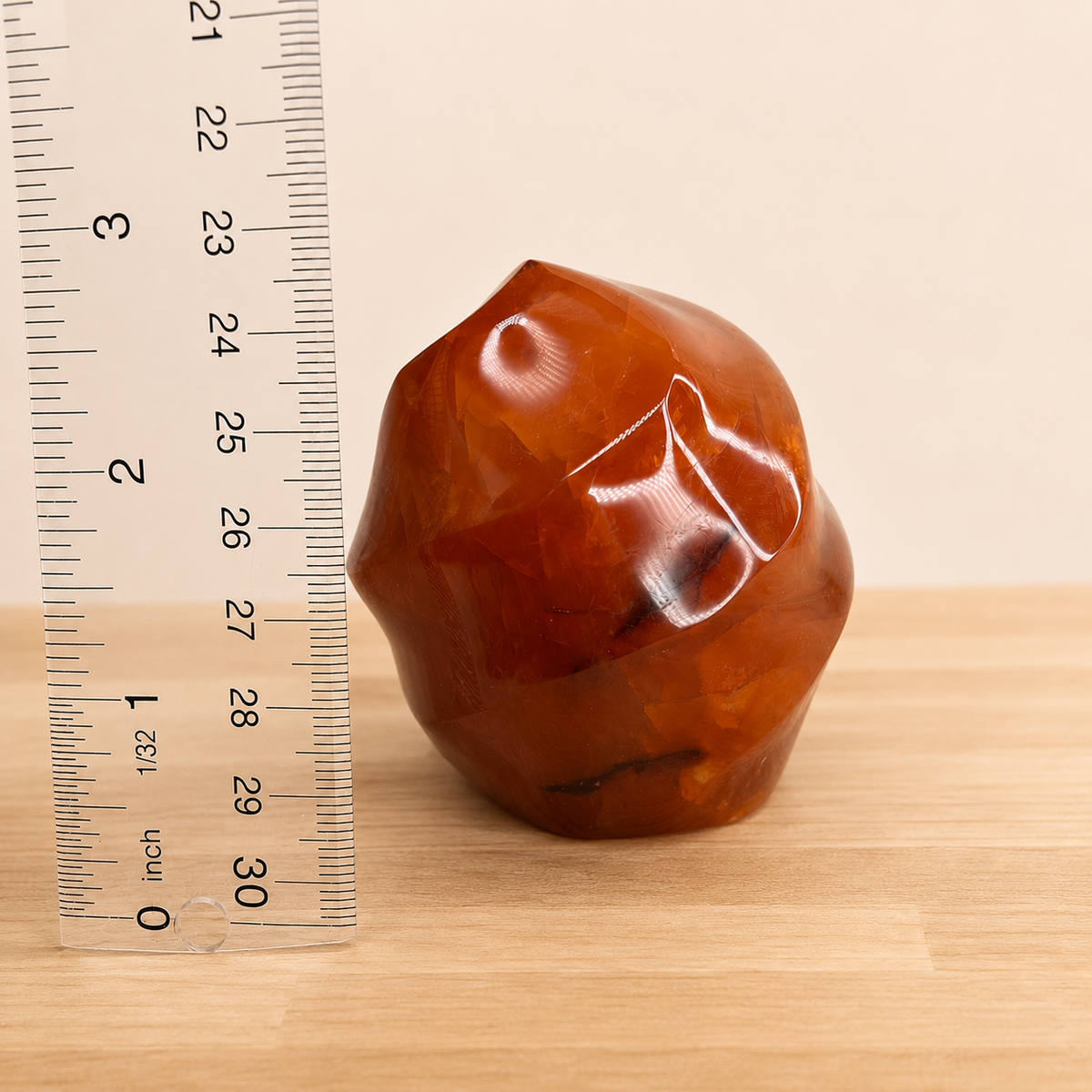 A polished brown gemstone or crystal with a faceted, organic shape sits on a light wood surface next to a clear ruler showing centimeter markings, against a plain beige background.