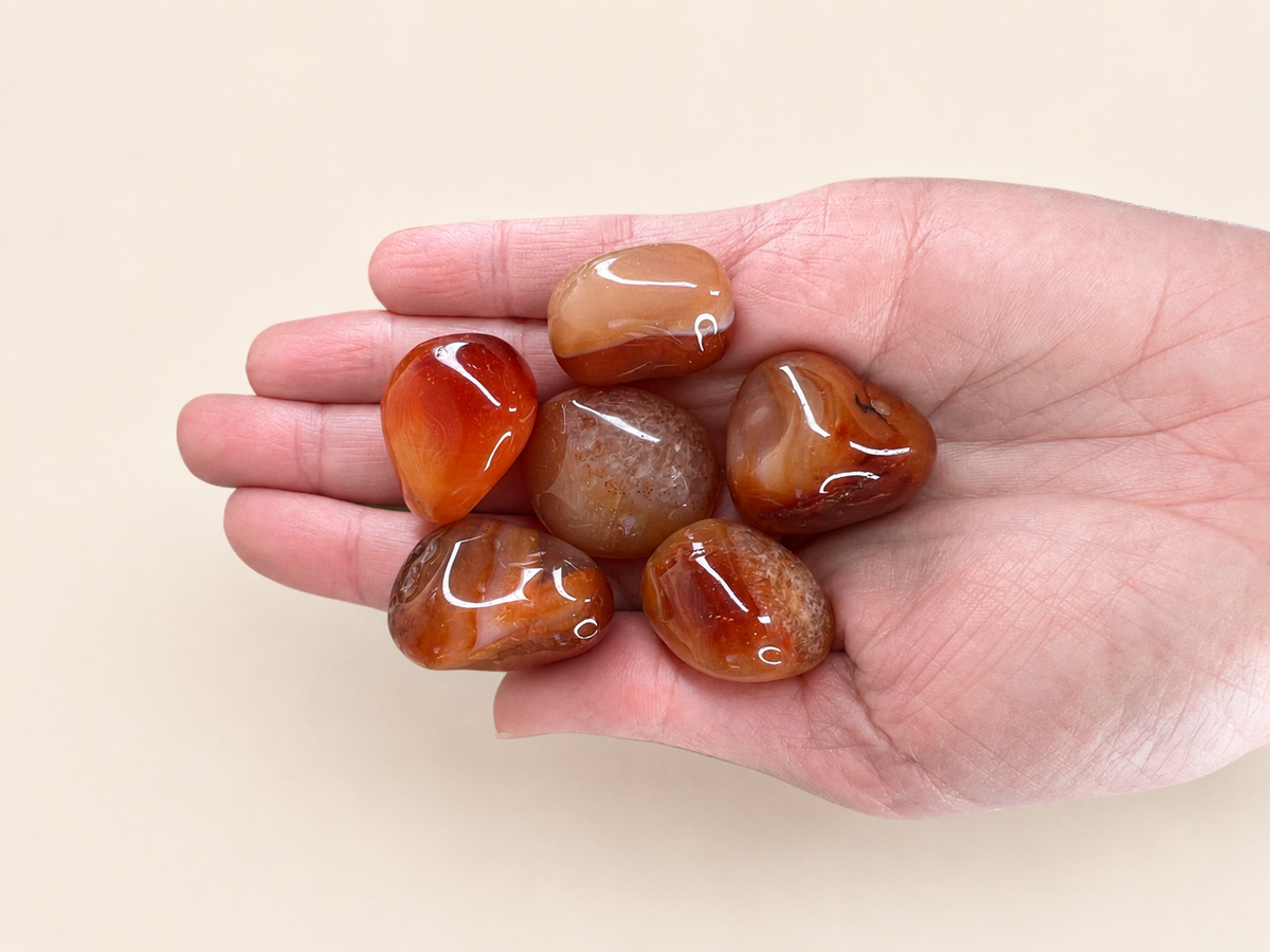 Five polished tumbled gemstones in warm orange, amber, and brown tones resting on an open hand against a plain beige background.