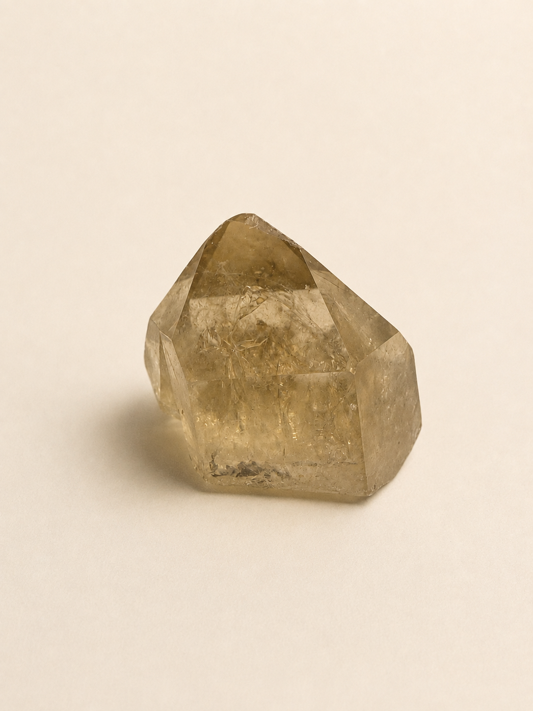 A natural faceted crystal stone with a pointed top, showing a translucent golden-brown color and internal inclusions, photographed on a plain light beige background.
