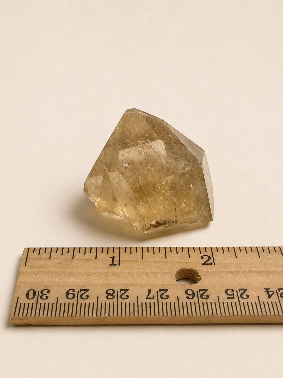A natural faceted crystal stone with a warm golden-brown hue, shown next to a wooden ruler for scale on a plain off-white background.