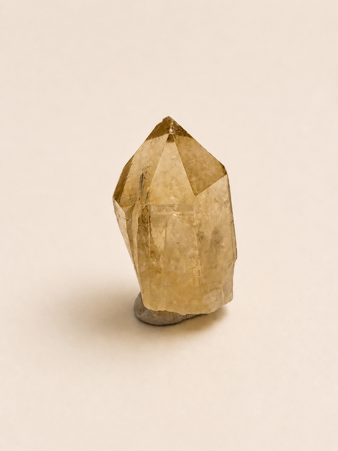 A natural yellow quartz crystal point with a polished geometric structure, resting on a small gray stone base against a solid, warm off-white background.