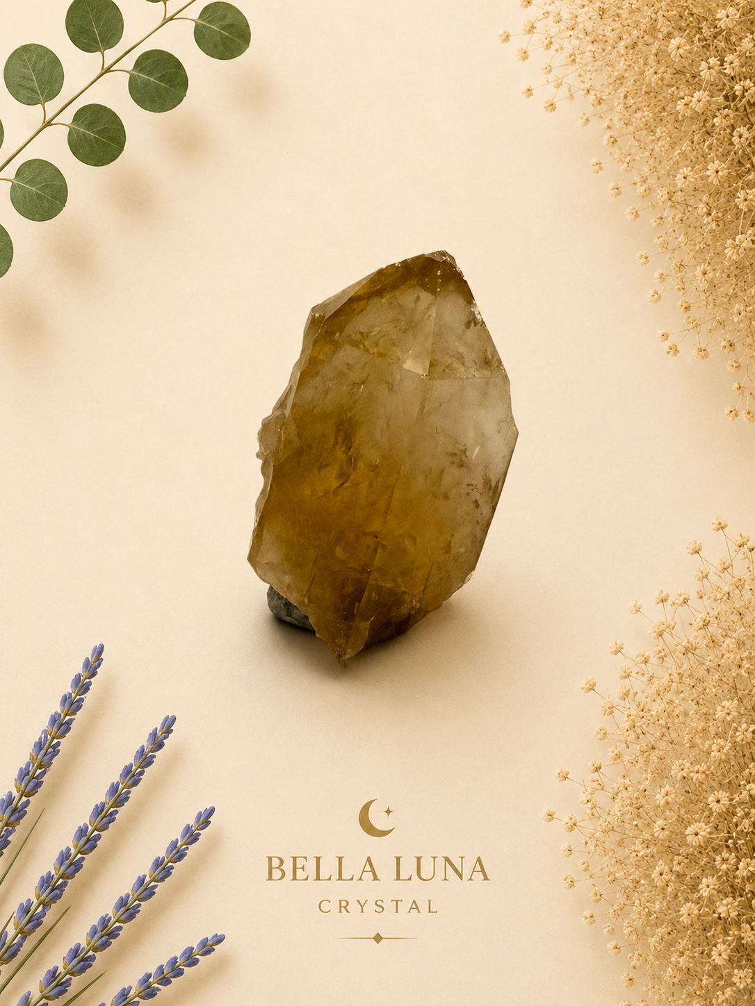 A raw crystal mineral specimen with a warm golden-brown hue, displayed on a light beige background with decorative botanical elements including lavender, eucalyptus, and gold glitter. The brand logo 'BELLA LUNA CRYSTAL' is printed at the bottom.