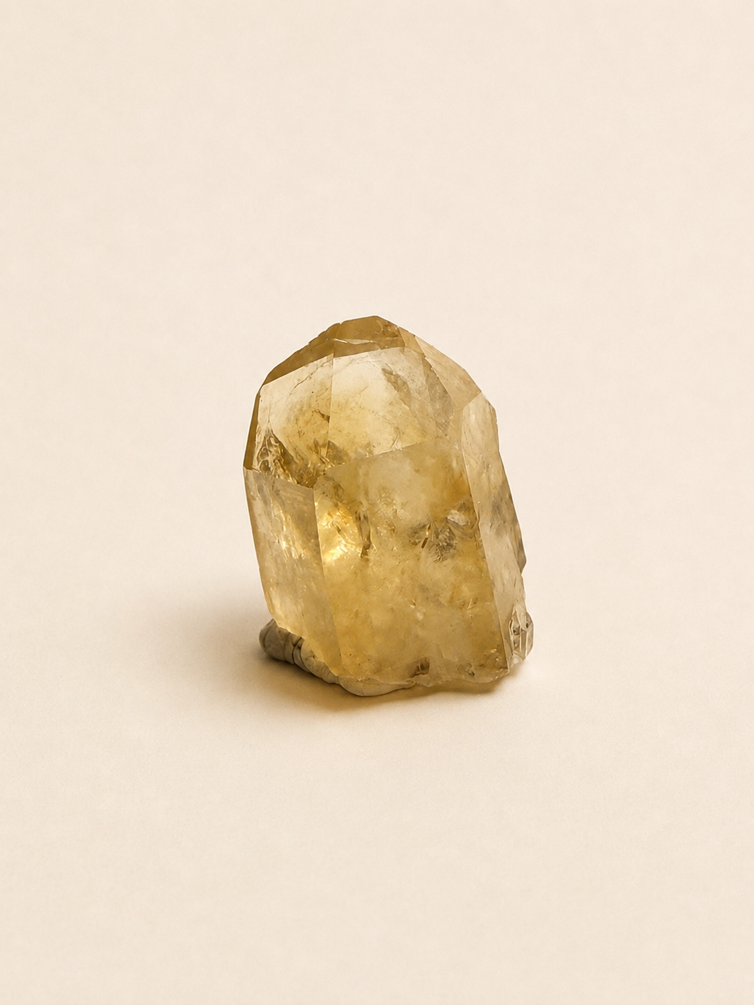 A raw, unpolished natural quartz crystal with a pointed tip and rough base, showing a translucent golden-yellow hue, set against a solid light beige background.