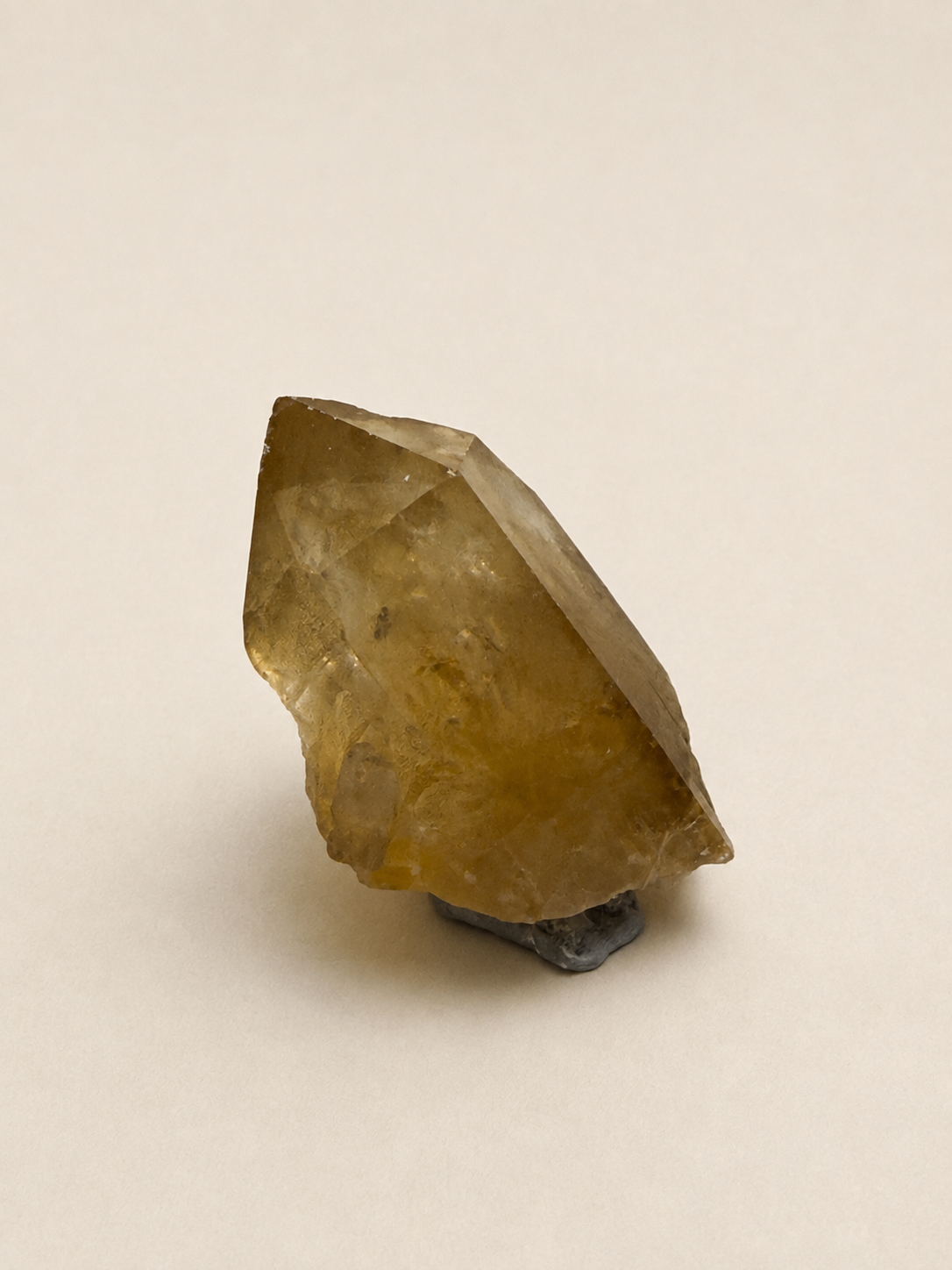 A raw, unpolished yellow-brown crystal mineral specimen resting on a small dark gray base, photographed against a plain, warm off-white background.