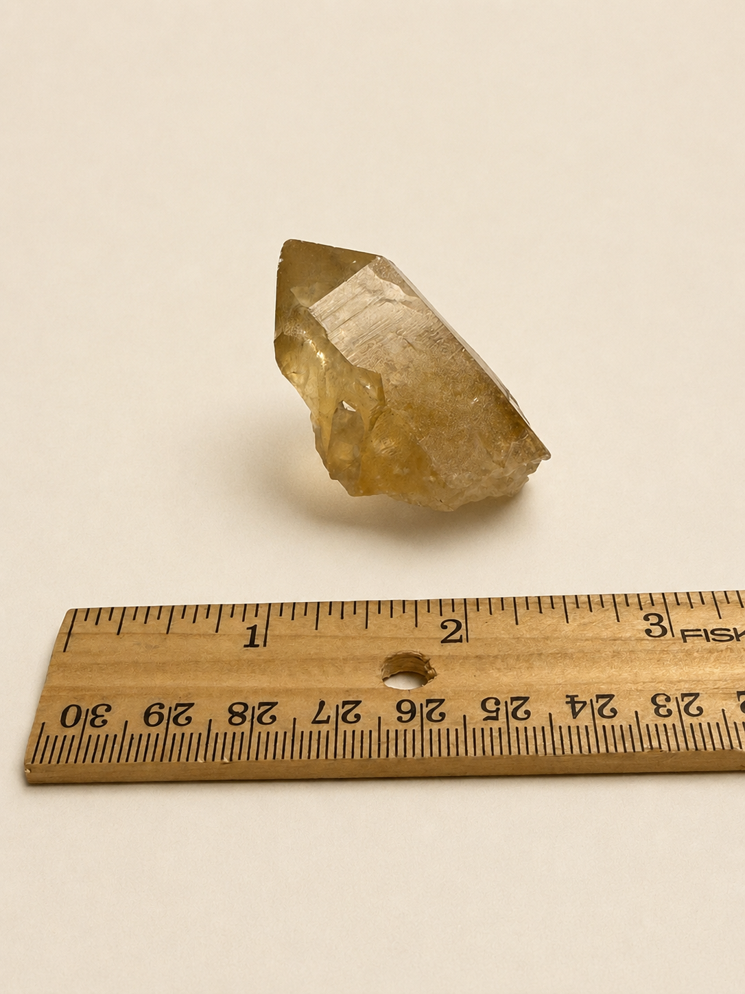 A rough, natural crystal or mineral specimen with a translucent golden-yellow hue, shown next to a wooden ruler for scale on a plain off-white background.