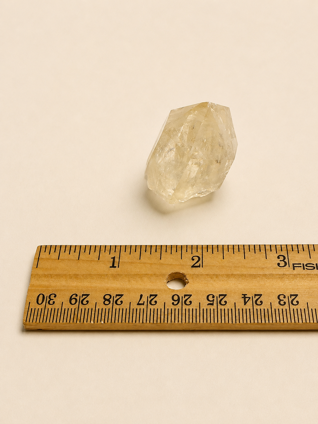 A small rough, pale yellow crystal or gemstone placed above a wooden ruler on a plain light beige background.