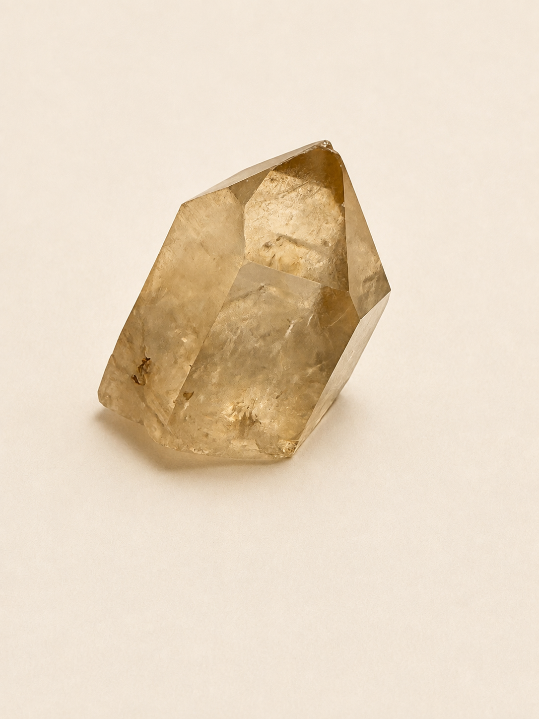 A faceted natural quartz crystal with a warm golden-brown hue, photographed on a plain, light beige background.