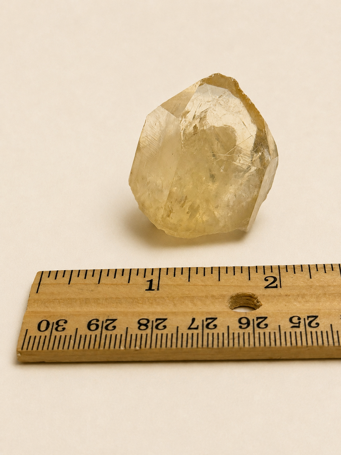 A faceted, rough semi-translucent yellow crystal stone shown above a wooden ruler for scale on a plain off-white background.