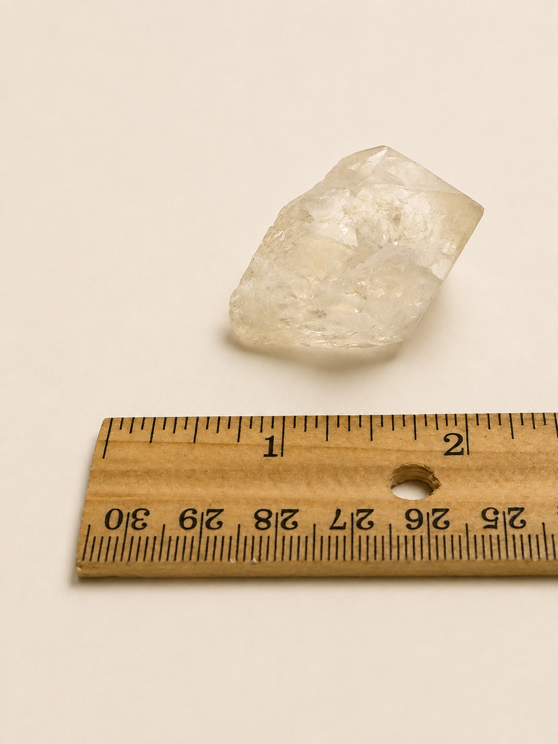 A rough, translucent pale crystal or gemstone placed above a wooden ruler on a plain light beige background.